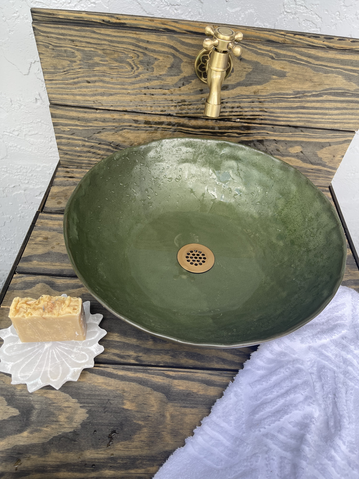 Green ceramic sink with gold faucet on a wooden surface with soap and towel.