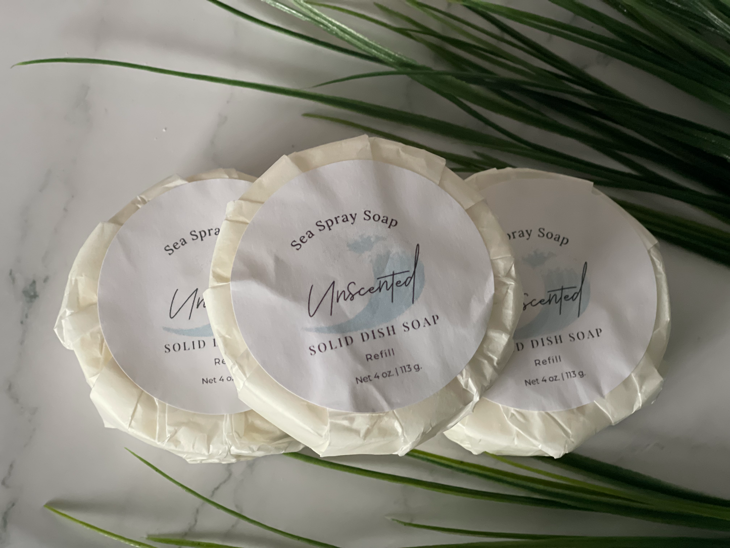 Three solid dish soaps with labels on a marble surface with green leaves.