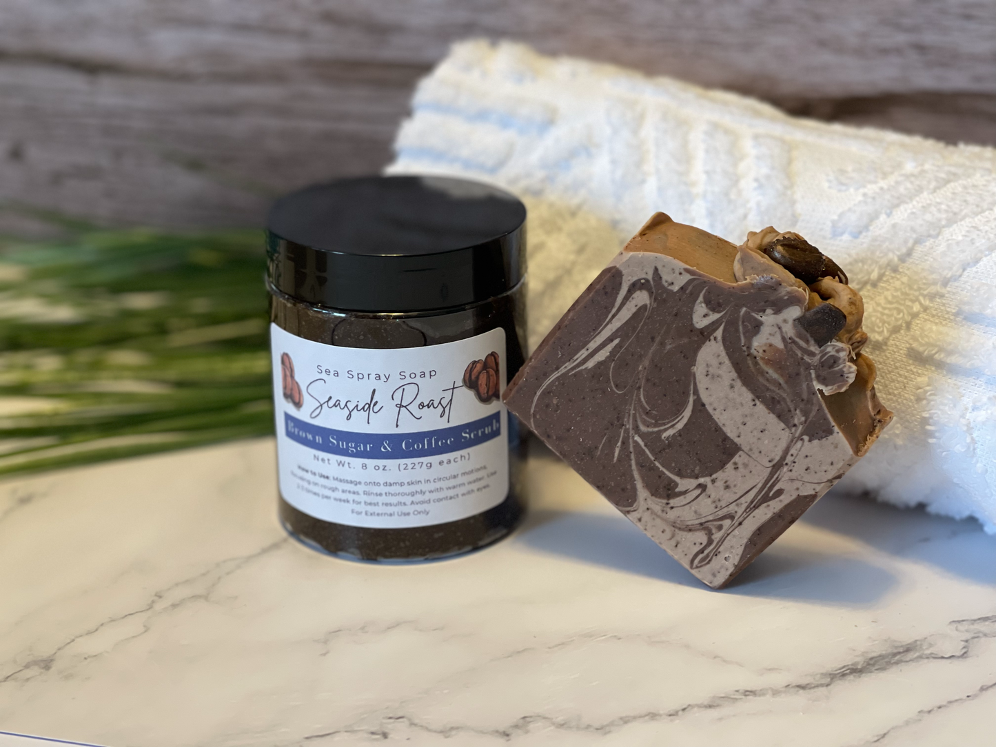 Jar of scrub and bar of soap on a marble surface with a towel and grass in the background