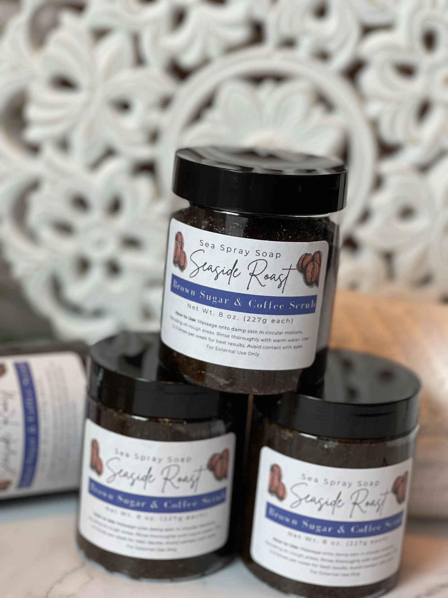Three jars of Seaside Roast Brown Sugar & Coffee Scrub stacked on a textured surface with a decorative wall in the background.