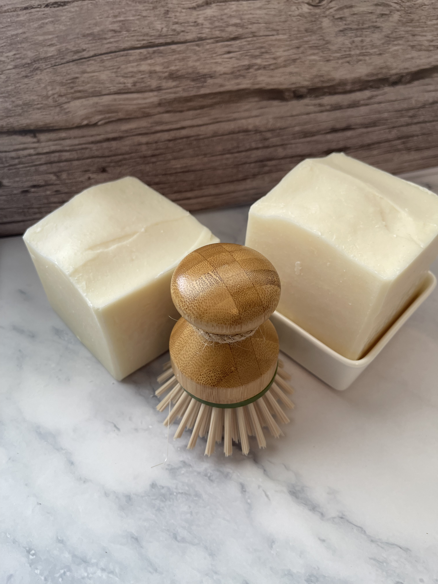 Two bars of soap and a wooden scrubber on a marble surface with a wooden background.