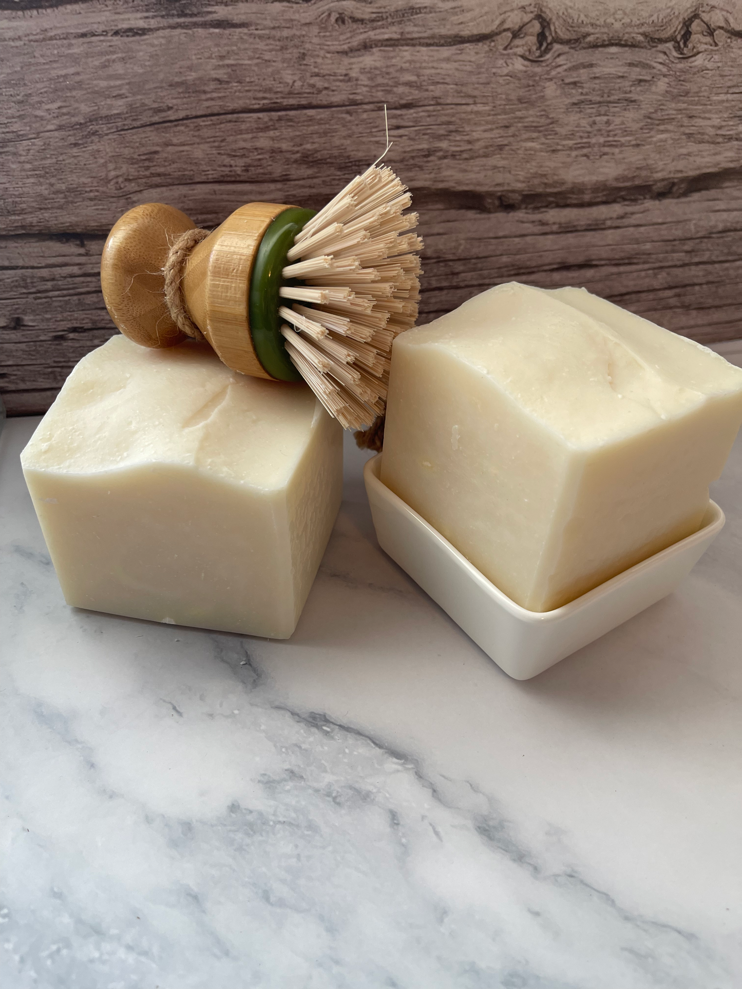 Two bars of soap on a marble surface with a wooden dish and brush in the background.