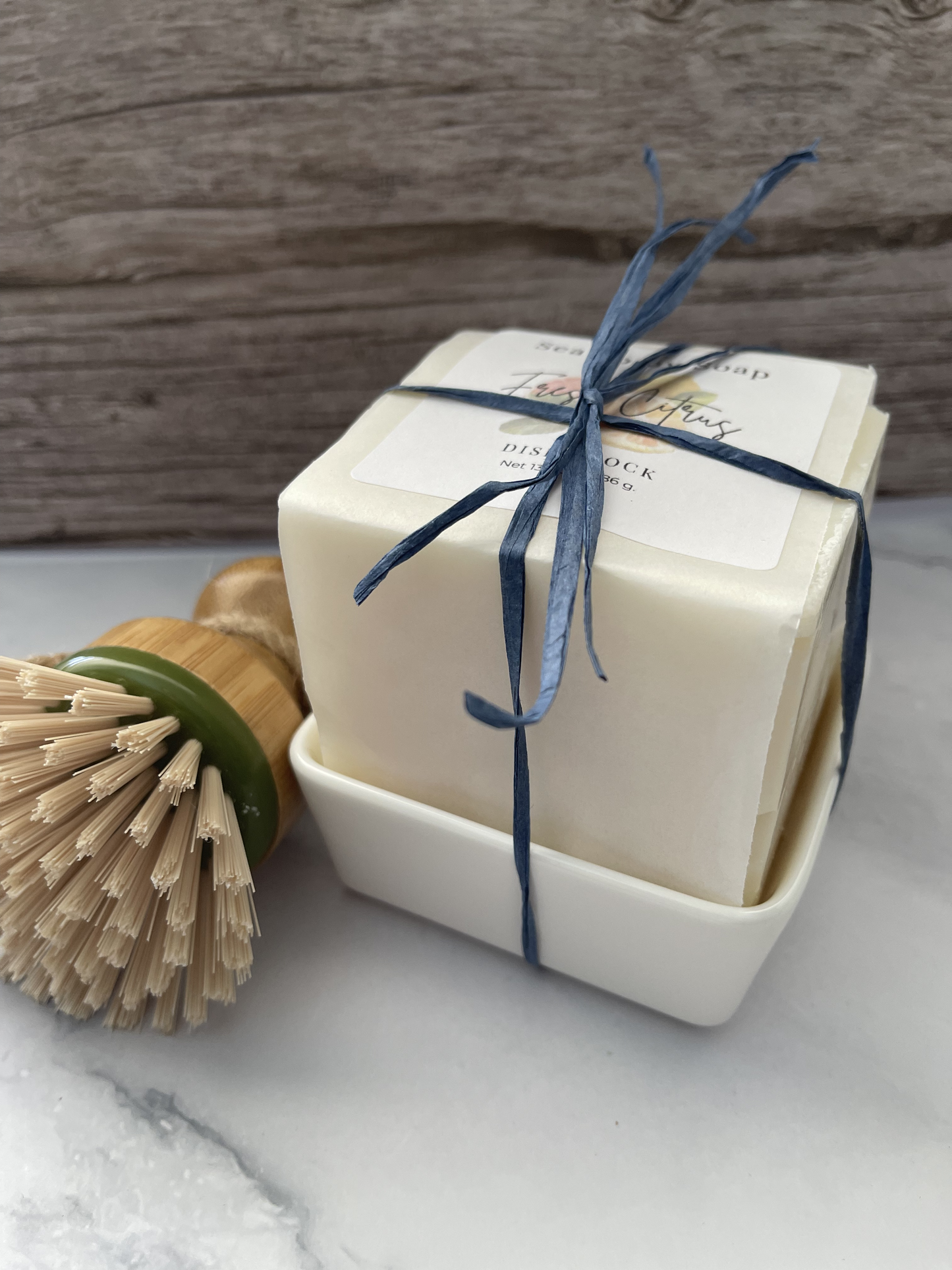 Bar of soap in a white box with a blue ribbon, next to a wooden brush on a light surface.