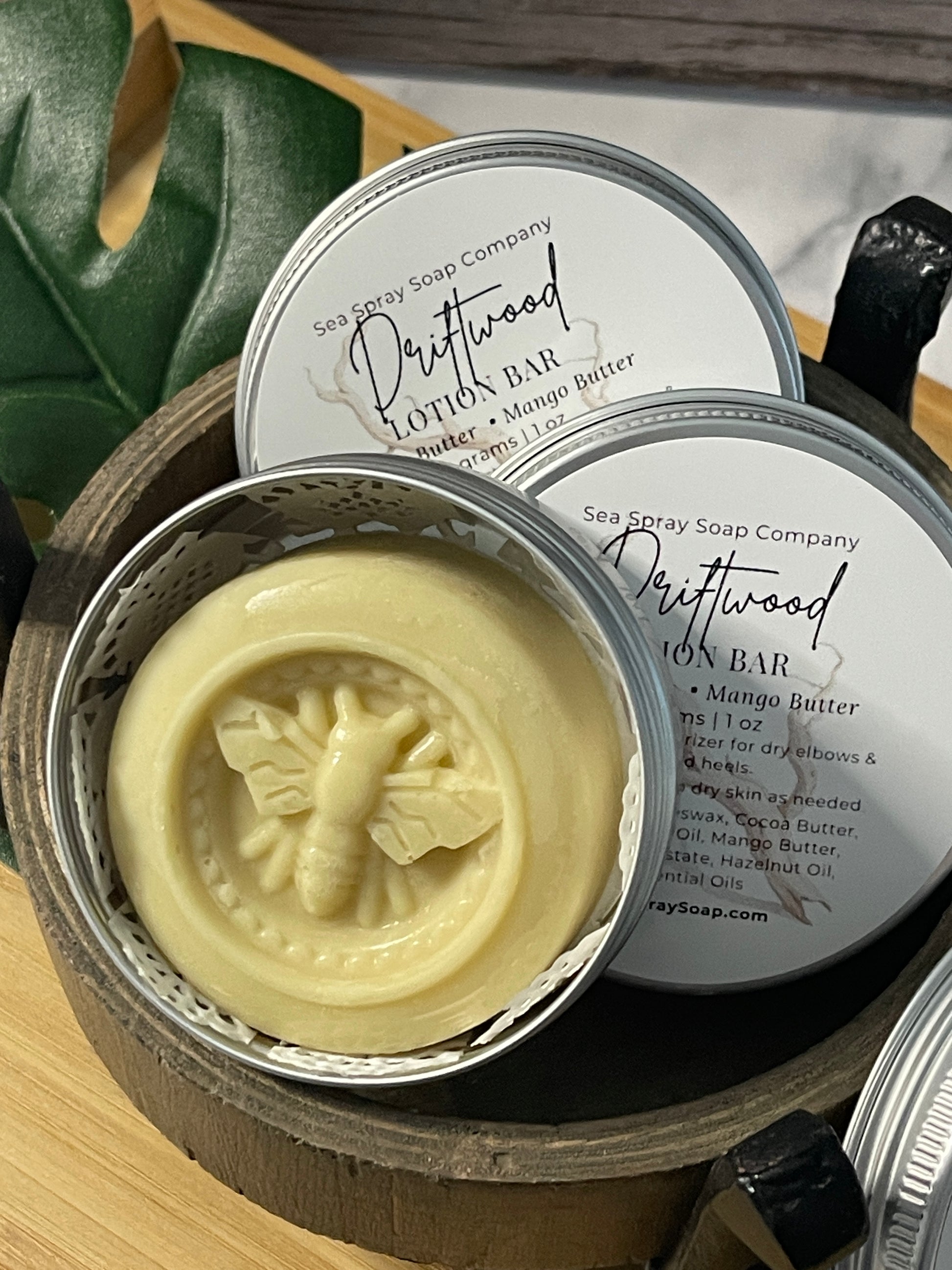 Bar of soap with a bee design in a metal container, surrounded by green leaves.