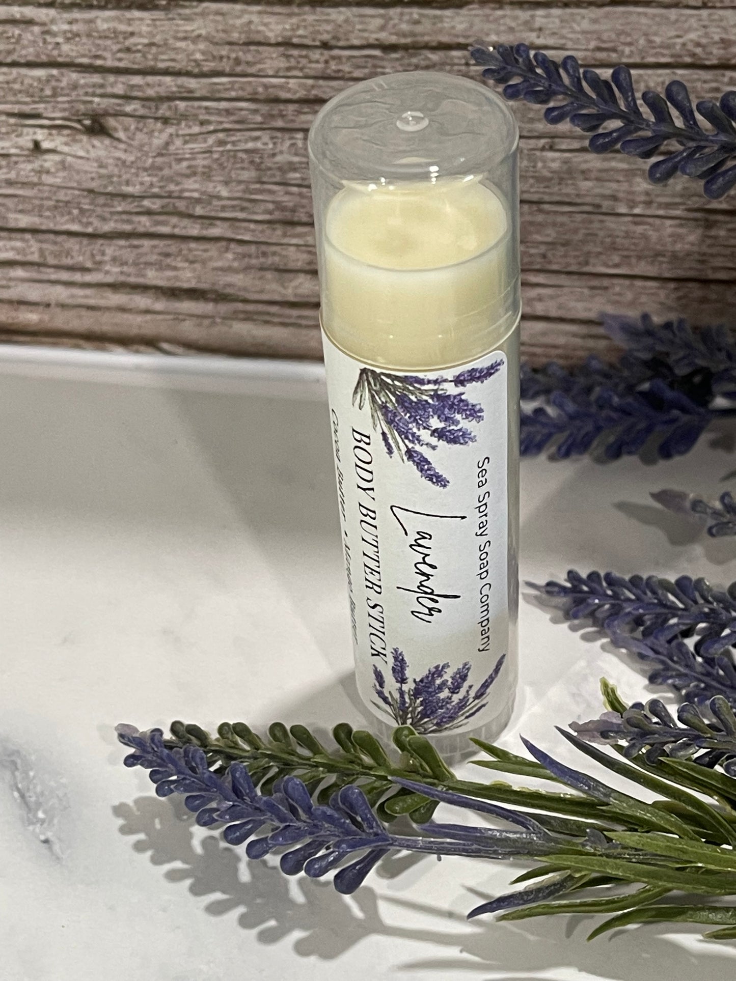 Lavender body butter stick with packaging on a wooden surface and lavender flowers.