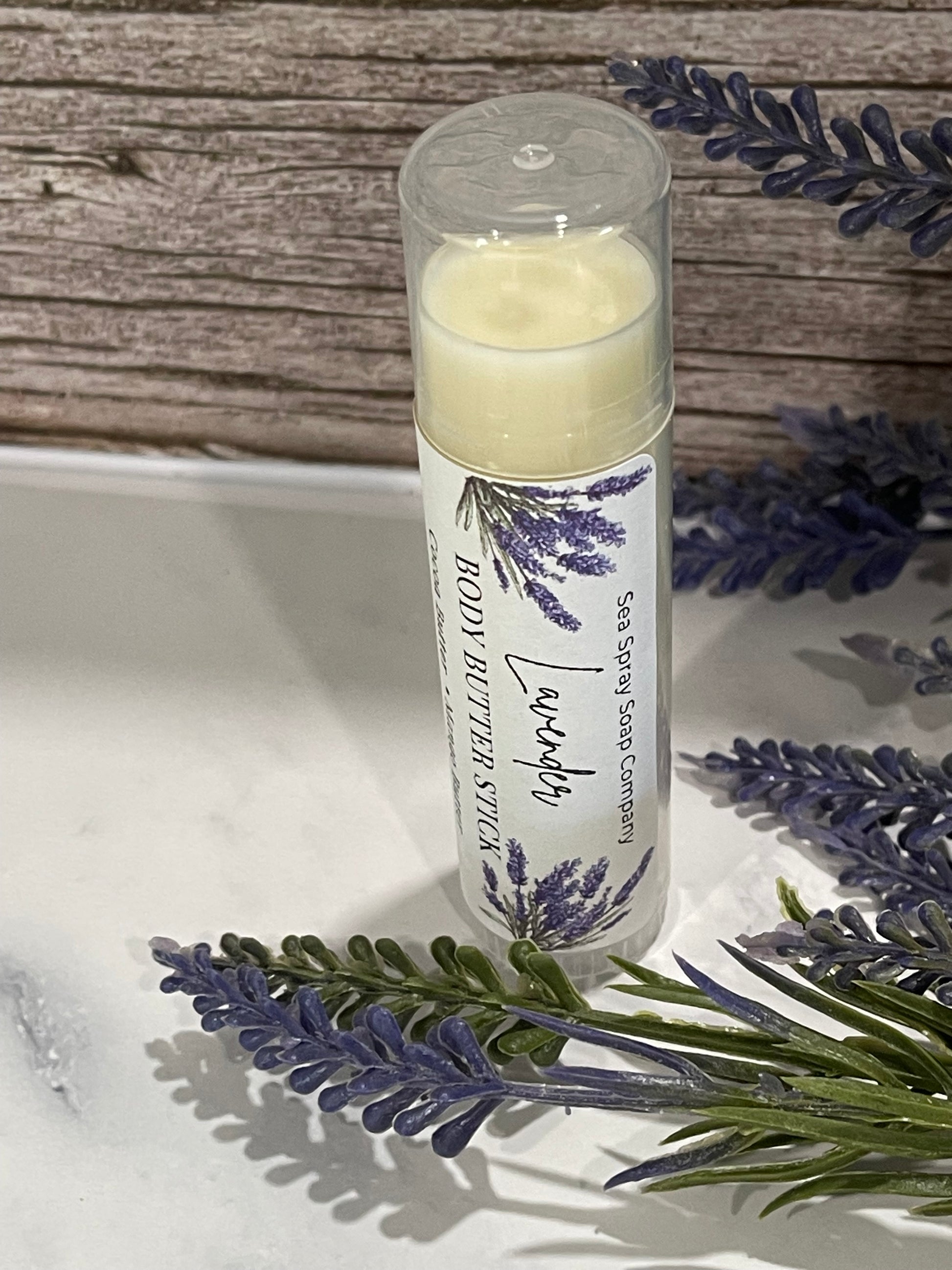 Lavender body butter stick with packaging on a wooden surface and lavender flowers.