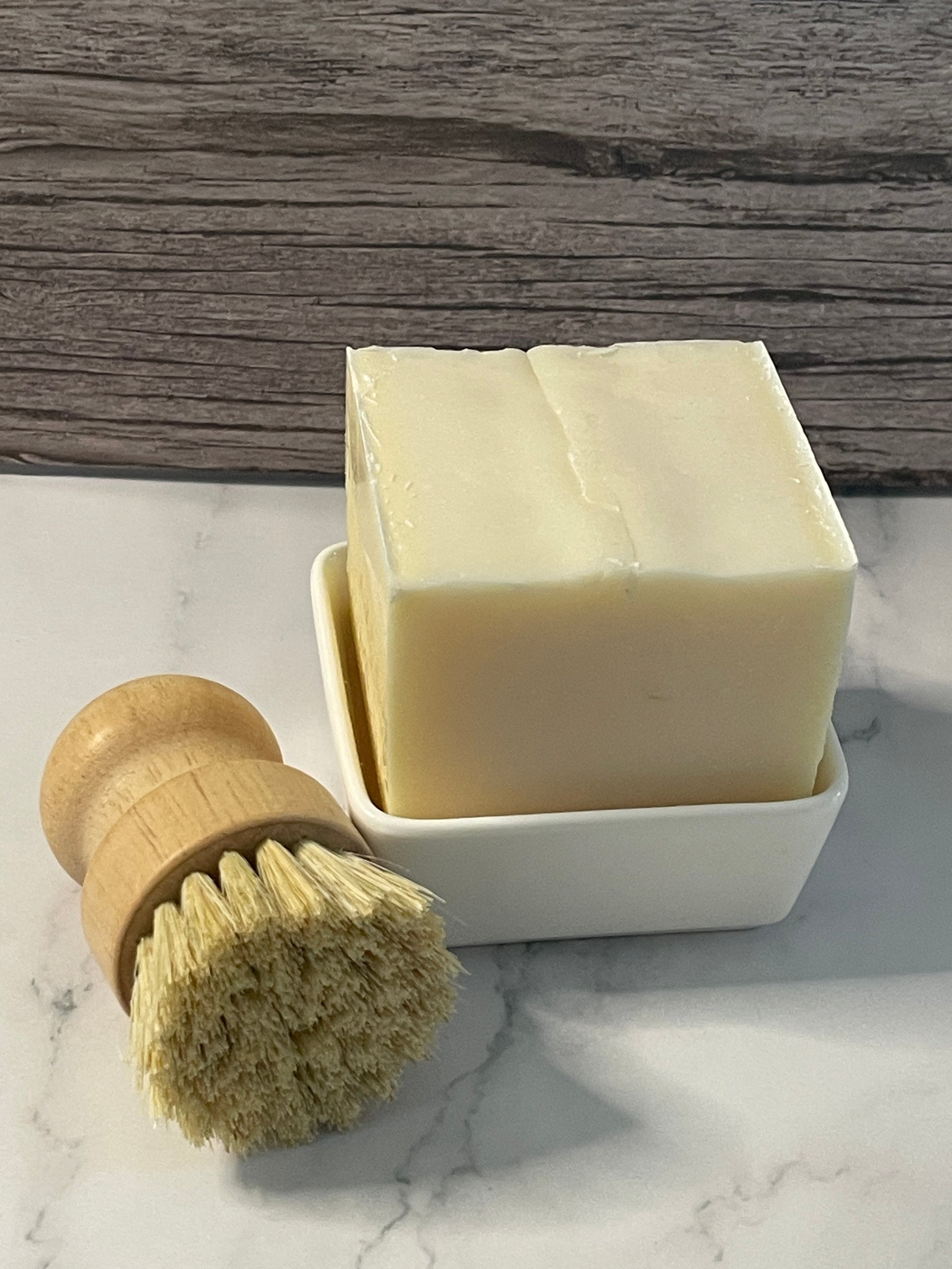 Bar of soap in a white dish with a natural bristle brush on a marble surface.