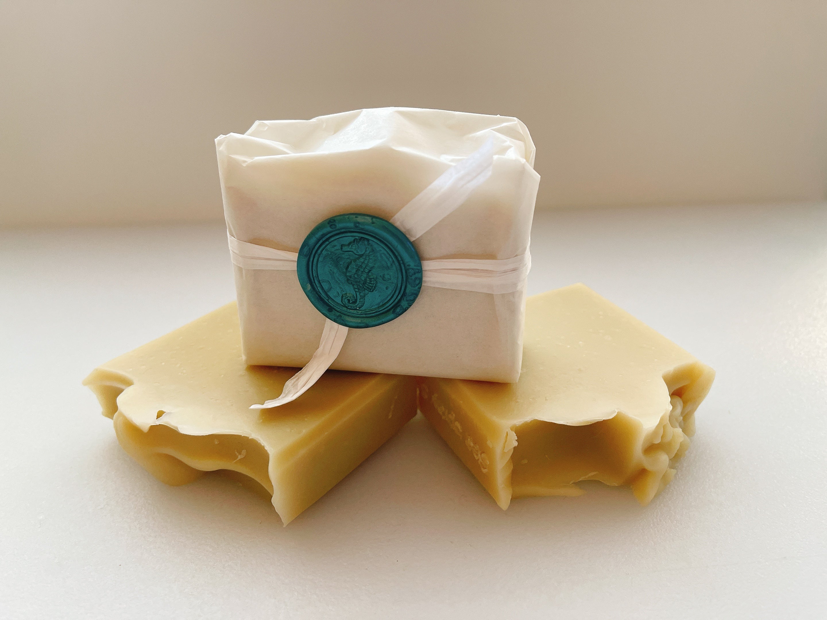 Two bars of soap with a wrapped bar on top, featuring a decorative seal, on a light background.