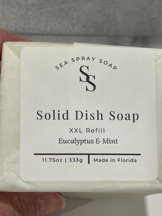 A hand holding a bar of Sea Spray Soap Solid Dish Soap XXL Refill in Eucalyptus & Mint scent, showing the white paper label with black text.