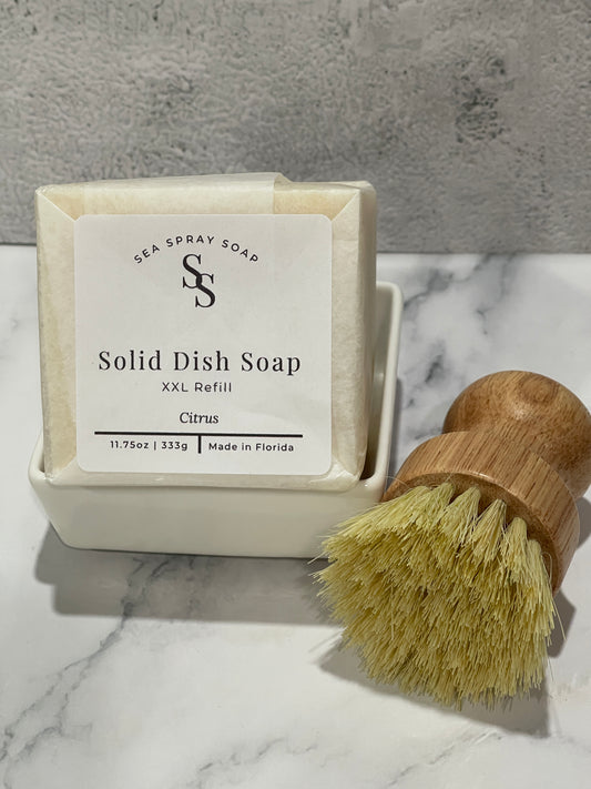 A packaged solid dish soap refill in a white dish soap holder next to a wooden-handled dish brush with pale yellow bristles on a marble surface.