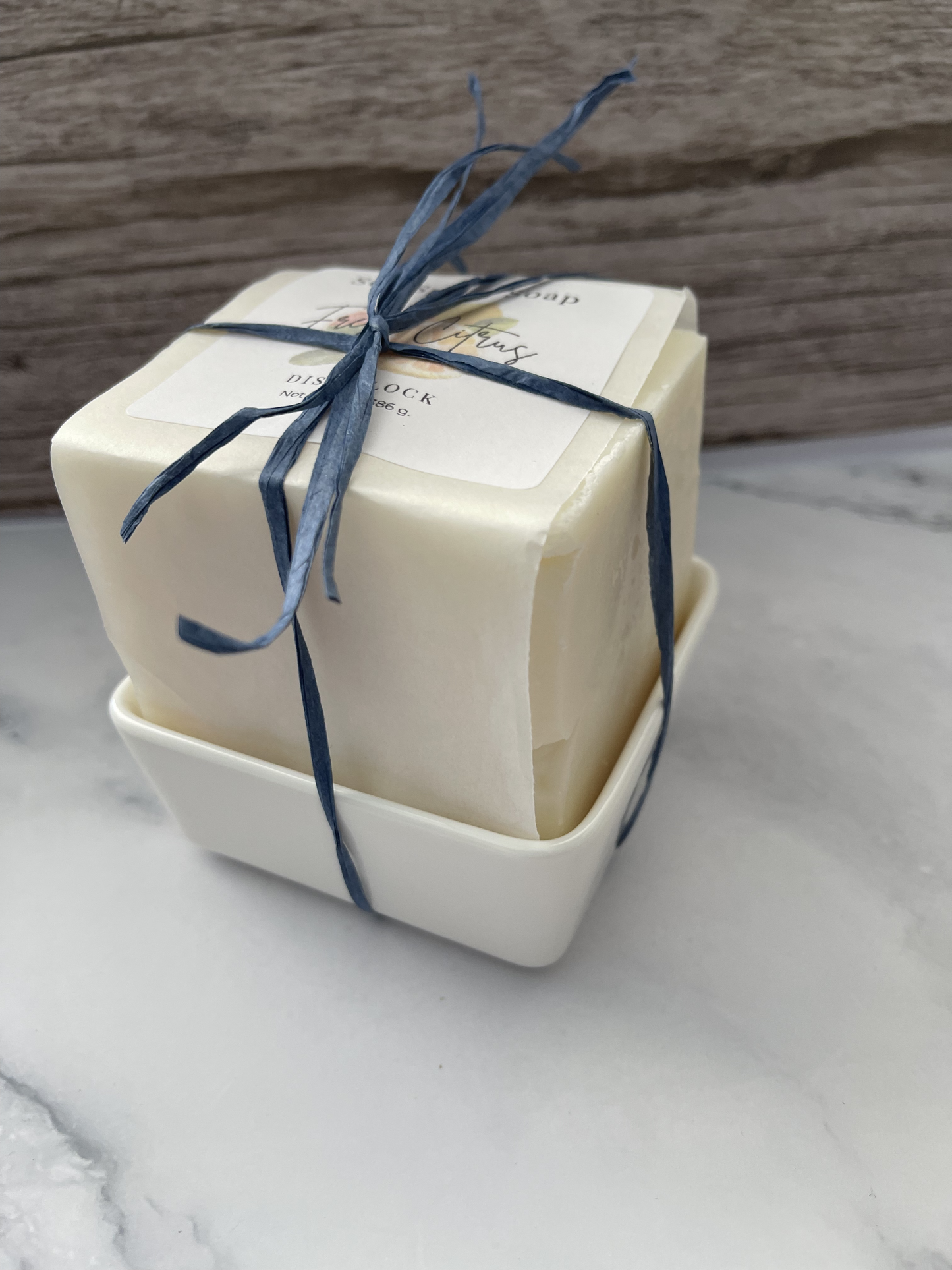 Two bars of soap in a white dish with blue ribbon on a marble surface.