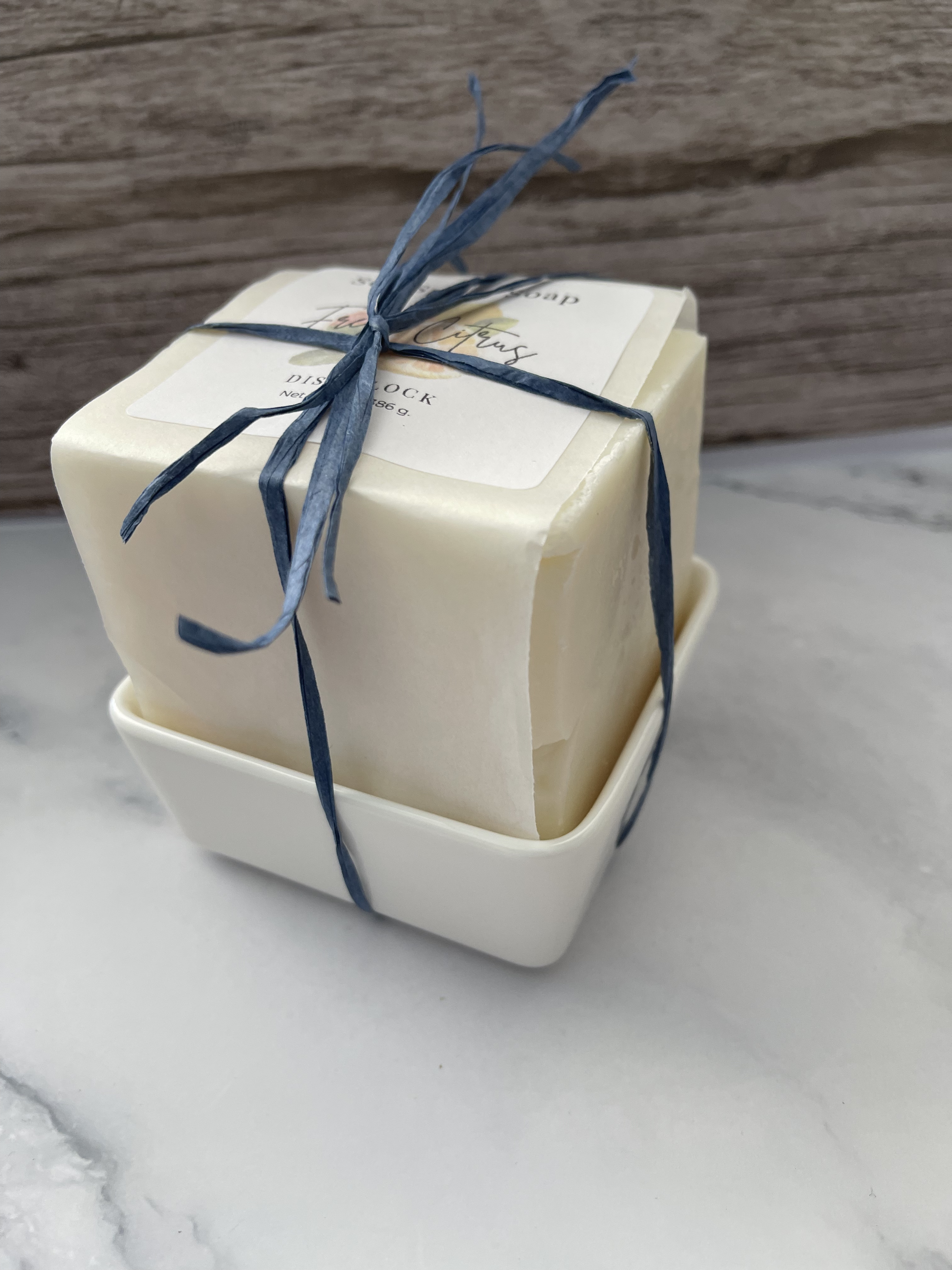 Two bars of soap in a white dish with blue ribbon on a marble surface.
