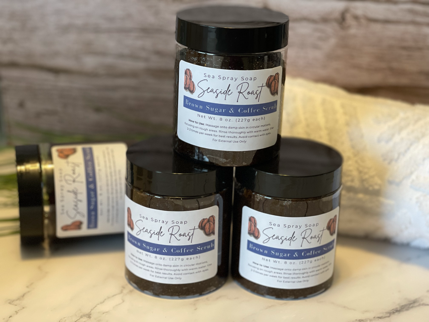 Three jars of Seaside Roast body scrub on a wooden surface with a blurred background.