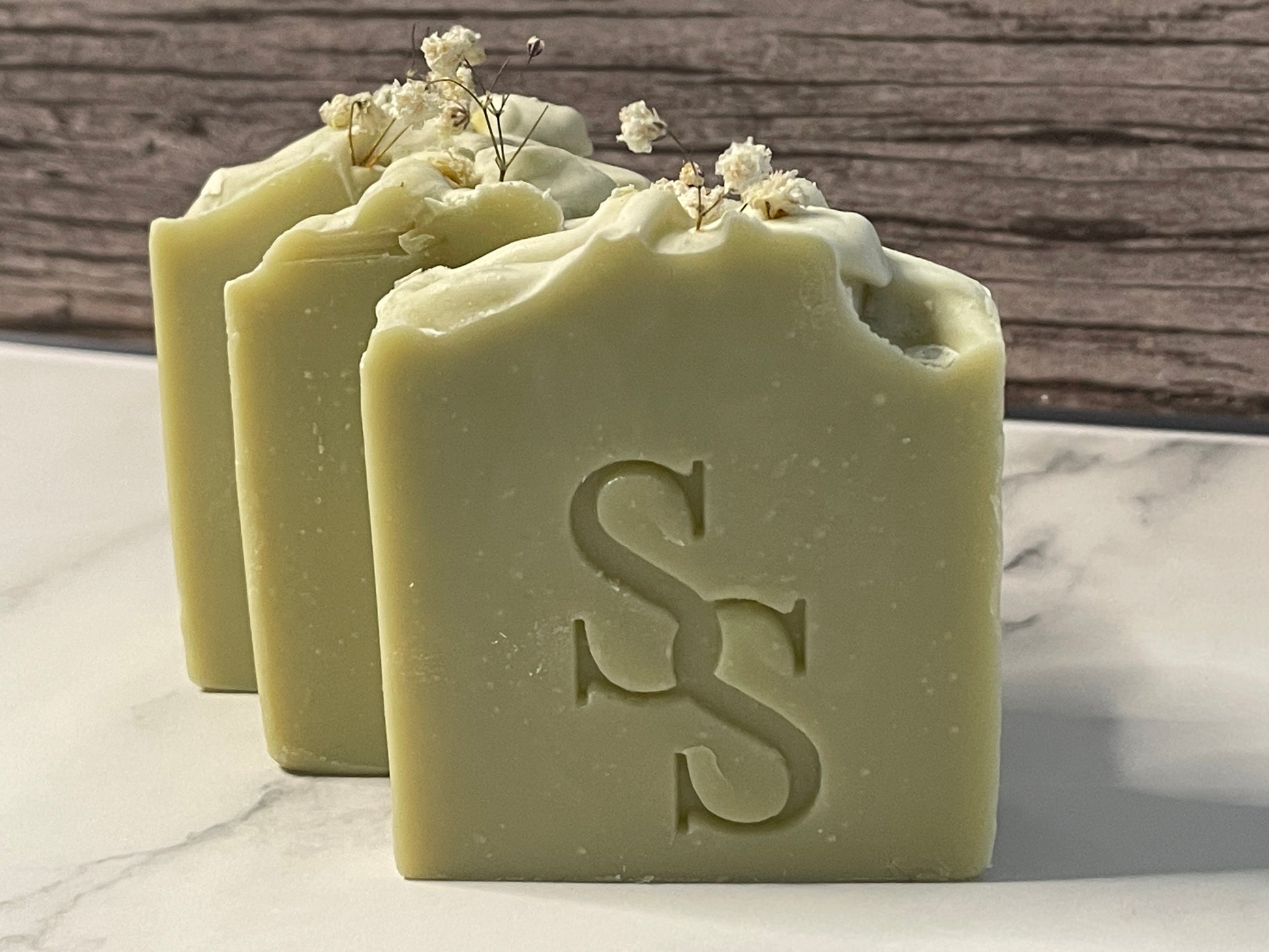 Three bars of green soap with a logo on a marble surface, surrounded by small white flowers.