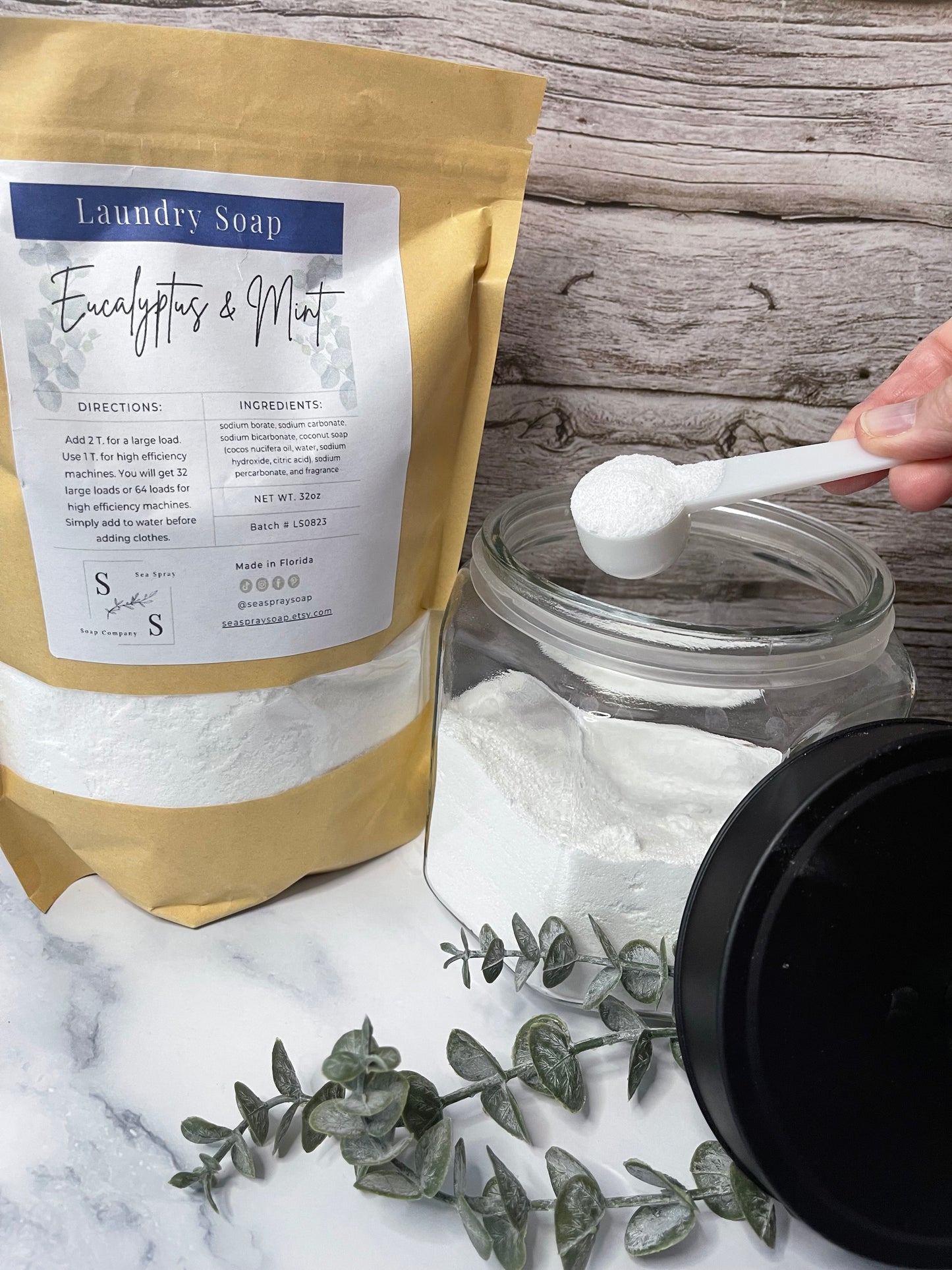 Person measuring laundry detergent with a spoon from a jar, next to a bag of detergent on a marble surface.