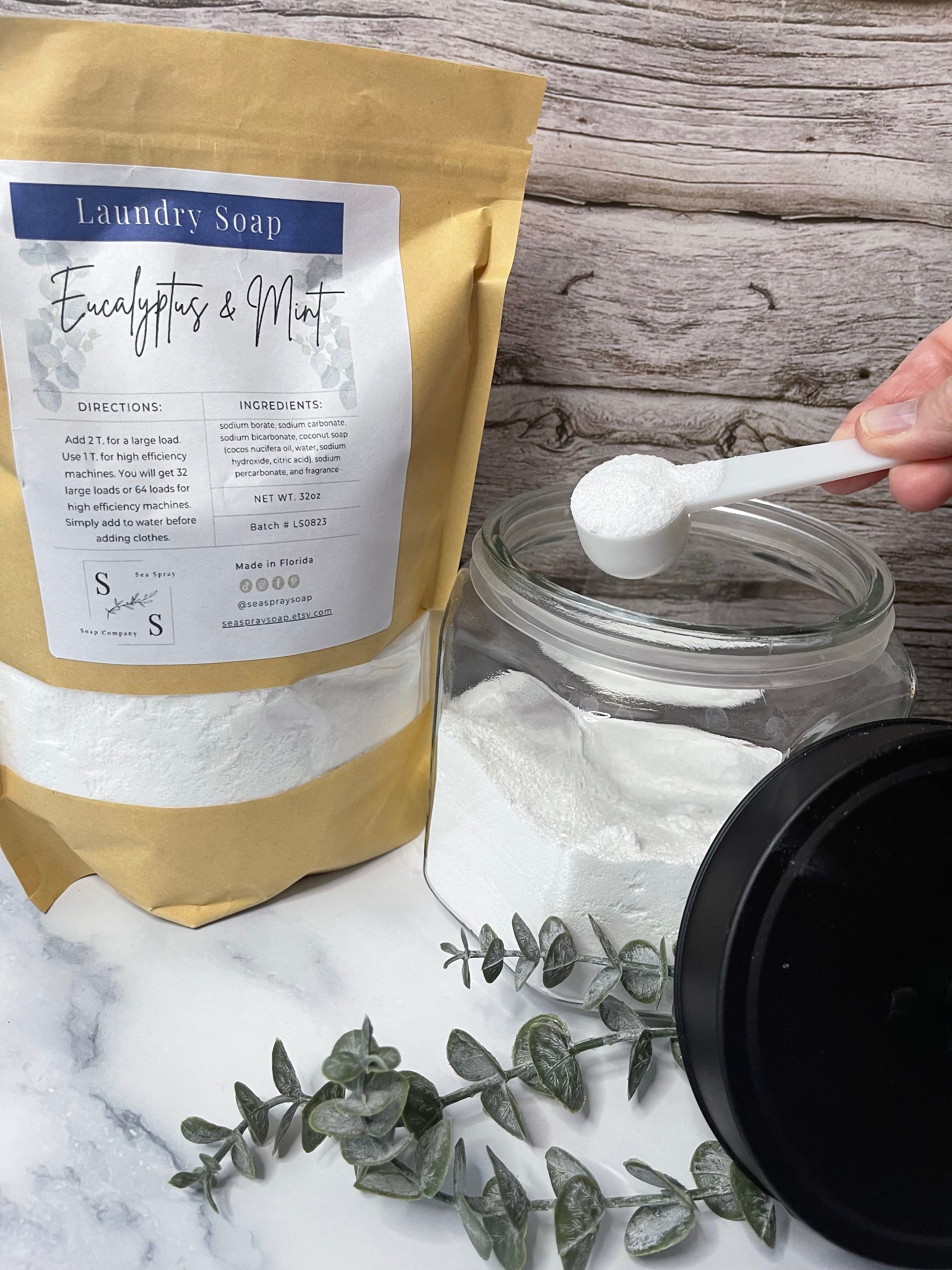 Person measuring laundry detergent with a spoon from a jar, next to a bag of detergent on a marble surface.