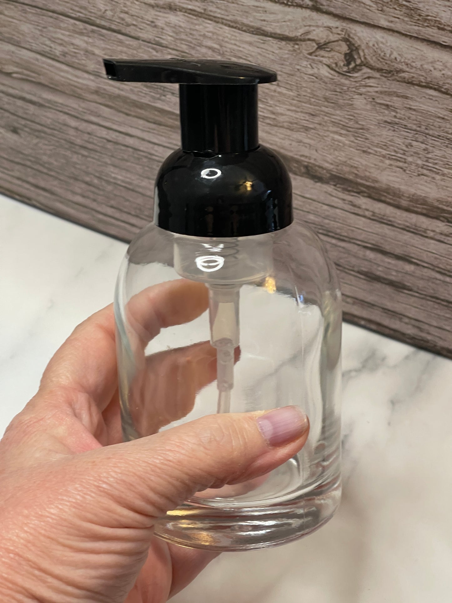 Hand holding a clear bottle with a black pump against a marble and wood background