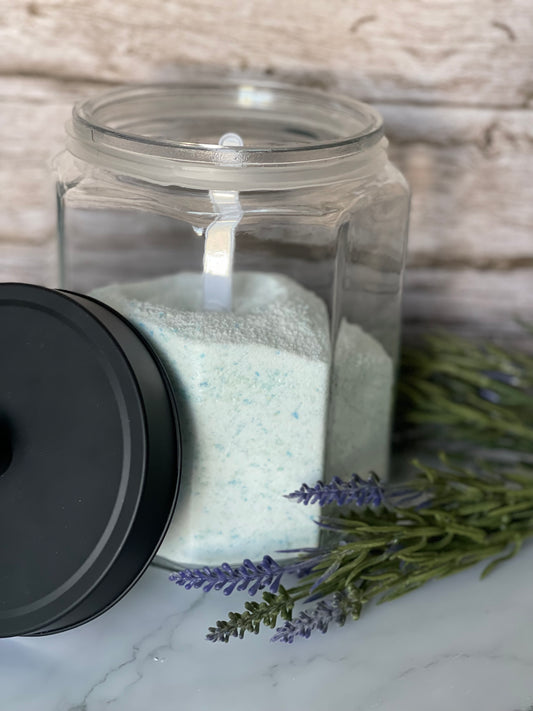 Clear jar with black lid containing a white substance, surrounded by lavender flowers on a marble surface.