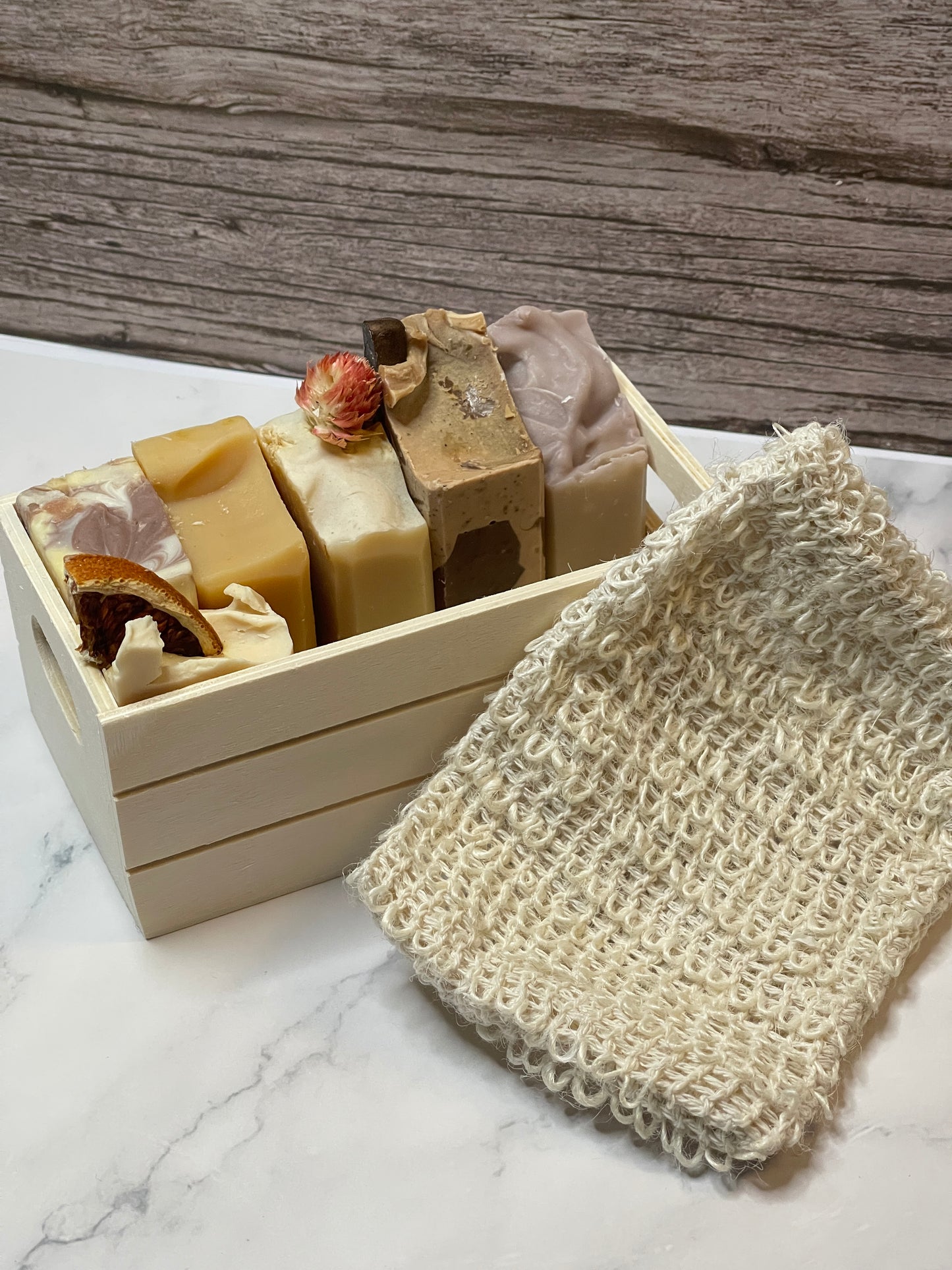 Six bars of soap in a wooden box with a loofah on a marble surface.