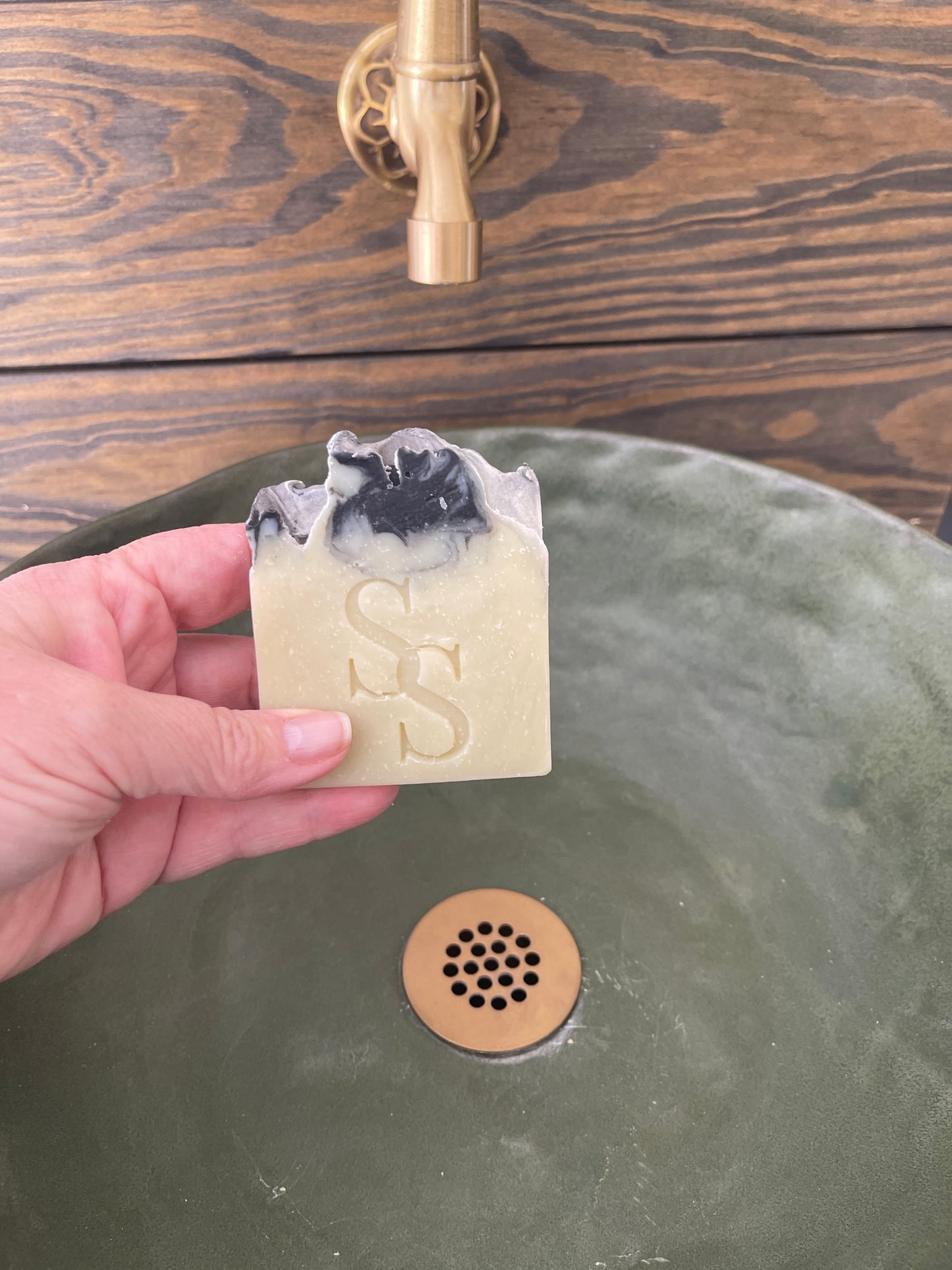 Hand holding a bar of soap with a brand logo next to a shower head and faucet.