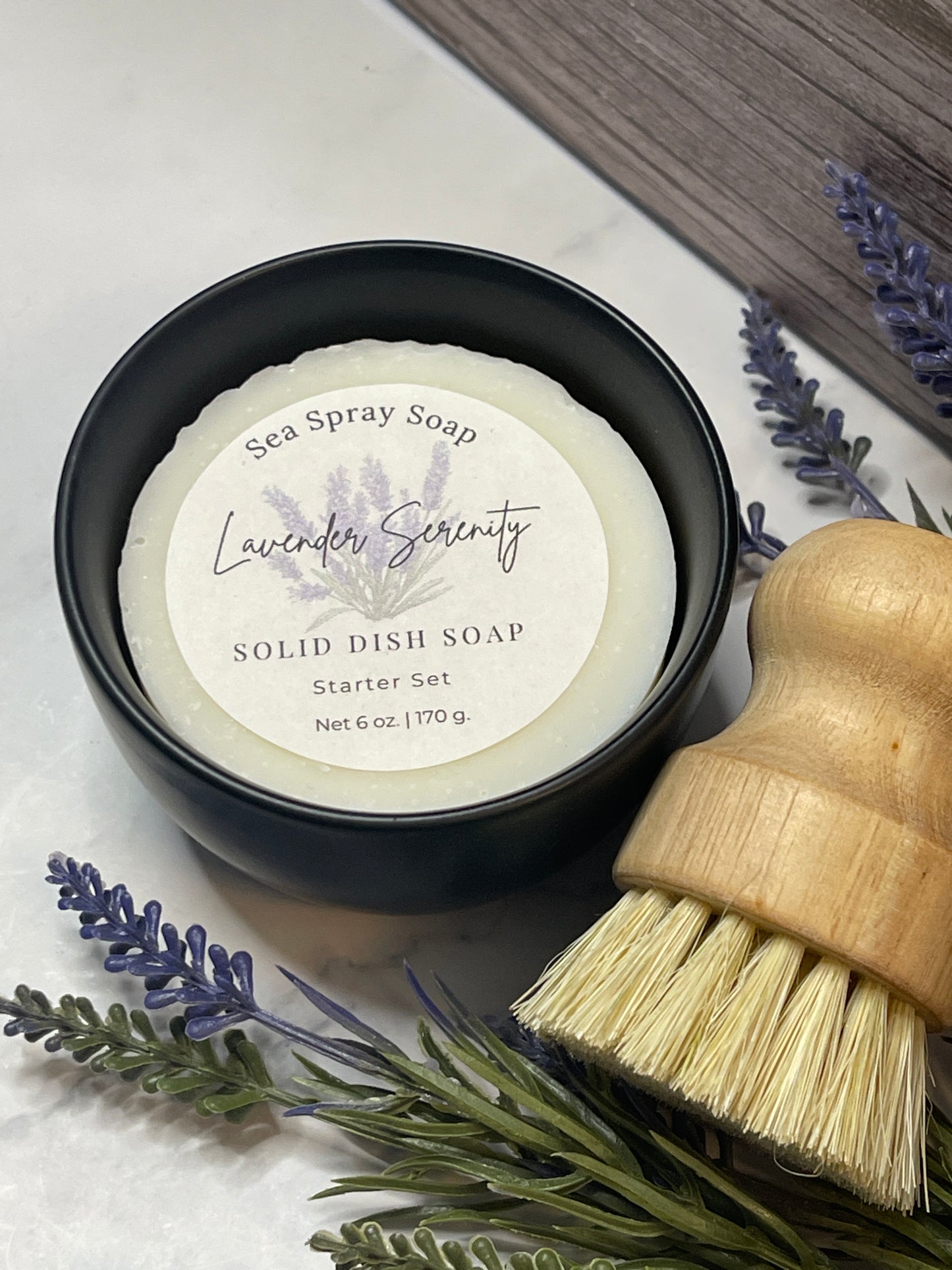 Solid dish soap in a black container with lavender sprigs on a white surface