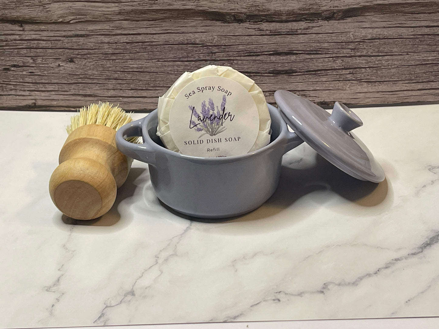 Solid dish soap bar in a gray container with a wooden brush on a marble surface.