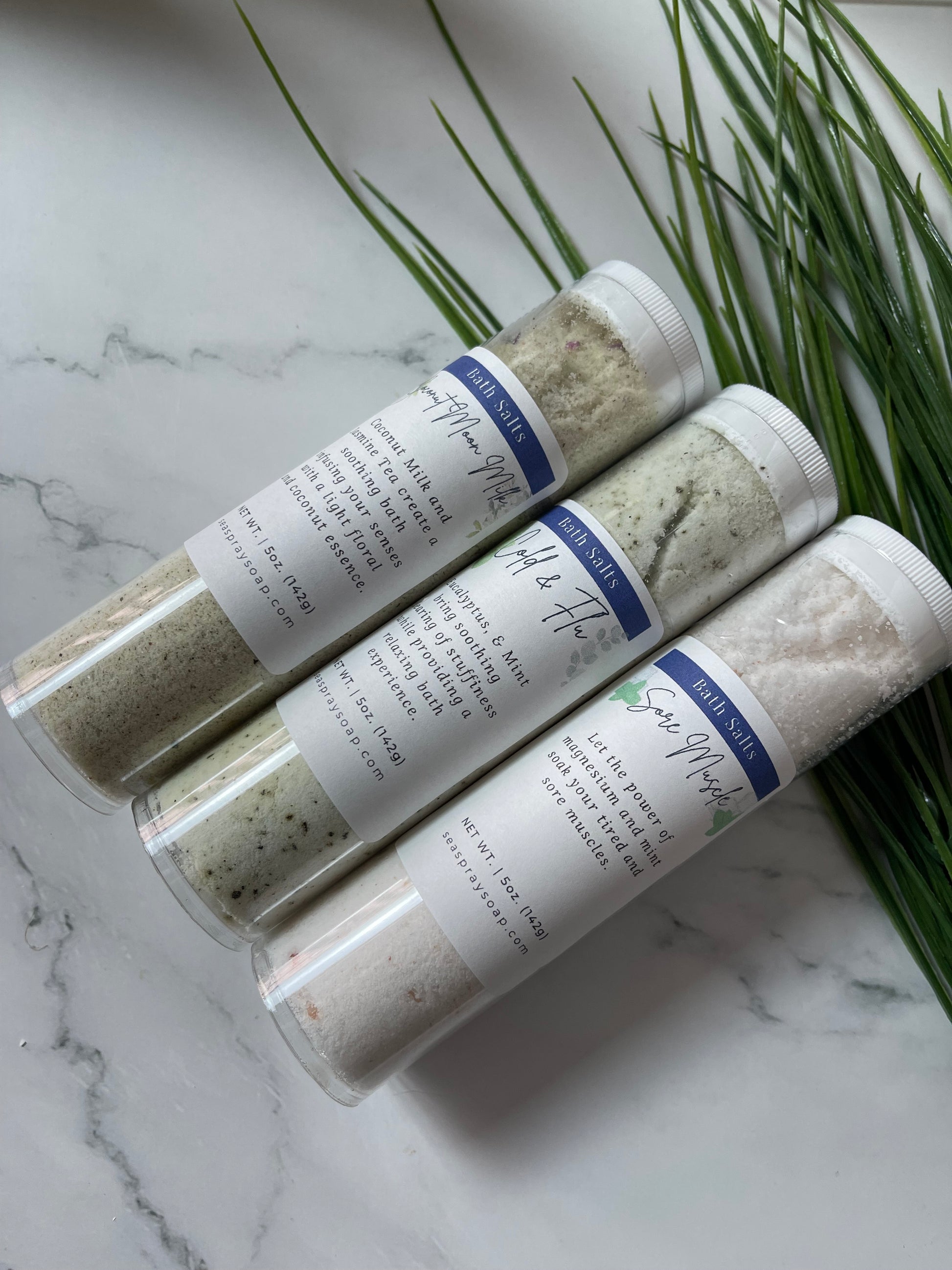 Three tubes of bath salts on a marble surface with greenery