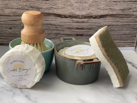Set of cleaning items including a dish brush, soap, and sponge on a marble surface with a wooden background.