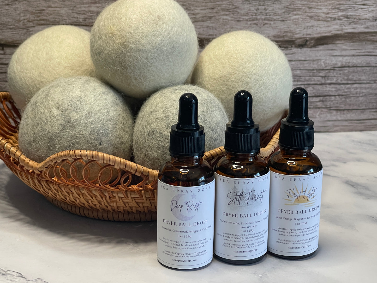 Three dropper bottles with labels next to a basket of dryer balls on a marble surface.