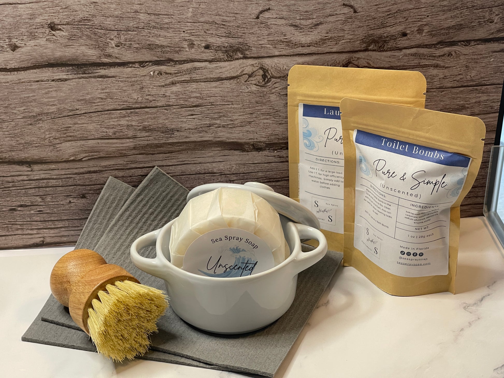 White ceramic dish for solid dish soap bar with a brush and two packages of unscented products on a wooden surface.