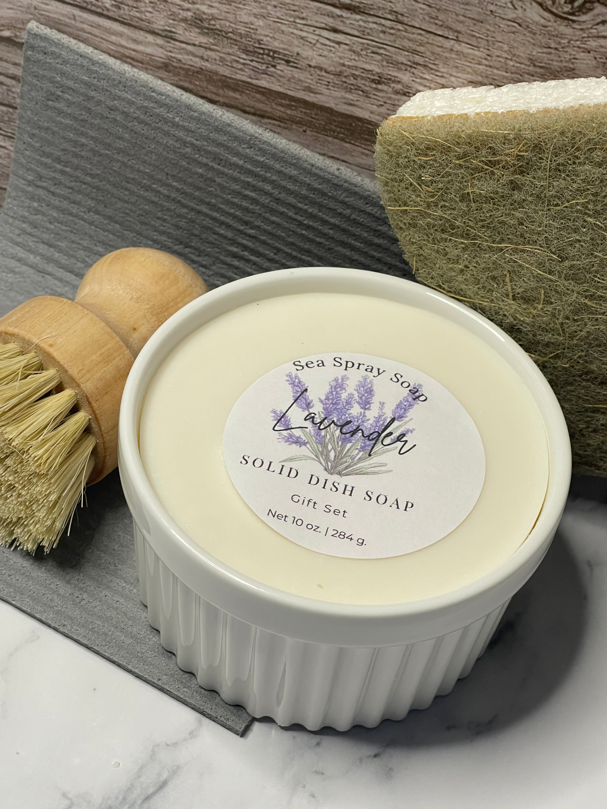 Solid dish soap bar with a label on top of a scrub brush and sponge on a wooden surface.